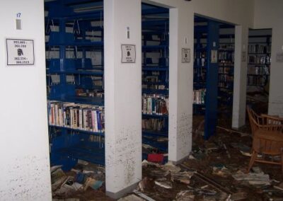A library shelving area with blue metal bookcases showing waterlines and missing items, while the floor is littered with mud, soaked books, and broken furniture. The space shows clear signs of storm surge destruction.
