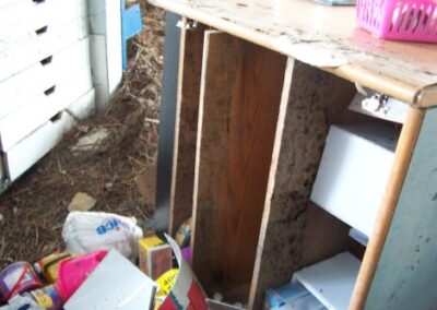 An open, damaged wooden desk shows missing drawers and mold‑stained interior surfaces, with debris and supplies scattered on the muddy floor. The top of the desk displays water damage and dirt.