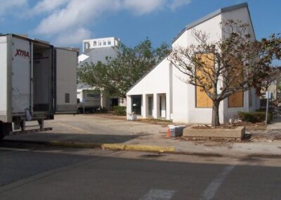 The exterior of the Biloxi Library shows boarded windows, scattered debris, and cleanup trucks parked nearby. A damaged tree stands beside the building along a debris‑strewn sidewalk.