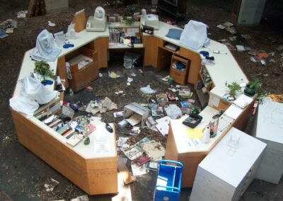 A circular reference desk is surrounded by scattered books, debris, mud, and damaged office supplies, with covered computer equipment resting on top of the counters. The floor inside and around the desk area is heavily littered with storm-damaged materials.