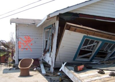 A collapsed white house leans off its foundation with broken walls and shattered windows, surrounded by scattered debris and a damaged wicker chair in the yard. Spray‑painted search‑and‑rescue markings are visible on the exterior.