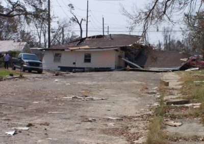A severely damaged house lies partially collapsed in the middle of a road, with debris covering the surrounding pavement. A vehicle is parked nearby as people inspect the storm destruction.