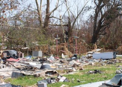 A grassy yard is covered with scattered storm debris, including broken appliances, metal fragments, insulation, and household items. Bare trees and snapped branches fill the background.