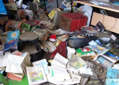 A heavily damaged library interior contains collapsed tables, scattered books, broken shelving, and warped ceiling tiles. A section of the cinderblock wall has fallen, exposing debris behind it.