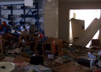 A library room filled with broken furniture, collapsed shelving, and piles of mud‑soaked books scattered across the floor. Light enters through a doorway, illuminating the extensive storm damage.