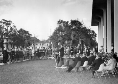 A large crowd gathered on the library lawn listens to a speaker at a podium, with rows of chairs set up for dignitaries and the building’s columned façade extending along the right side.
