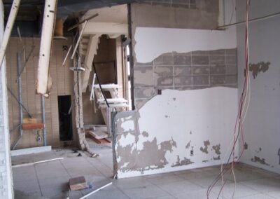 A damaged room inside the Gulfport Library shows partially stripped walls, exposed cinderblock, and broken ceiling pieces hanging overhead. Debris and construction material are scattered across the tiled floor.