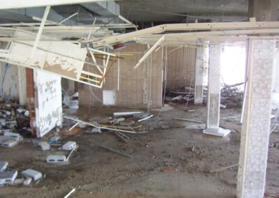 A large open section of the Gulfport Library interior shows collapsed ceiling pieces, exposed support beams, and debris scattered across the dirt‑covered floor. Several wall sections remain standing but show clear signs of flood and structural damage.