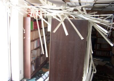A library shelving area shows books still on the shelves while broken ceiling panels and hanging metal strips cascade downward. The floor below is littered with debris and soaked materials.
