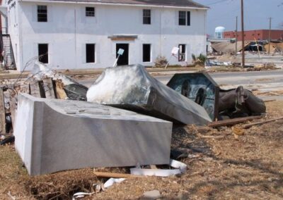 Broken pedestal blocks and the toppled bronze statue lie scattered along a roadside surrounded by storm wreckage near the Gulfport Library. A damaged white building stands in the background amid debris.