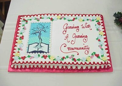 A decorated sheet cake features a drawing of a tree growing from an open book with the words “Growing With A Growing Community” written beside it. The cake is bordered with colorful icing flowers.