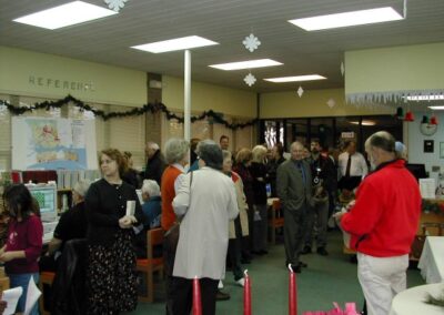 A crowd of people inside a library decorated with holiday garlands and hanging snowflakes gathers near bookshelves and service desks. Guests mingle and converse during a community event.