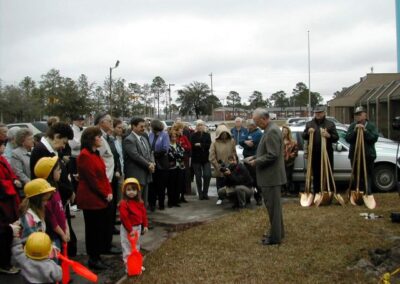 A man addresses a crowd at an outdoor gathering near a patch of grass while several gold ceremonial shovels stand upright nearby. Children wearing yellow hard hats and holding toy shovels stand at the front of the audience.