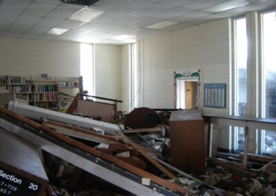A room inside the Pass Christian Library is filled with collapsed shelving, fallen furniture, scattered books, and water‑damaged debris. Light filters through tall windows illuminating the destruction.