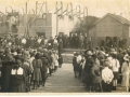A historical photograph showing a large crowd gathered in two long lines along a wooden walkway, with buildings and additional people visible in the background.