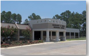 Exterior view of a two‑story brick library building with tall white columns at the entrance, large vertical windows, and a sign in front that reads “Harrison County Library System.”