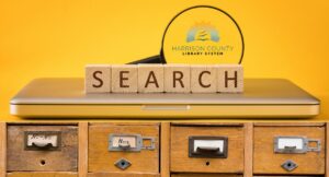 Wooden blocks spelling “SEARCH” on a laptop, with a magnifying glass showing the Harrison County Library System logo, and a row of vintage card catalog drawers below.